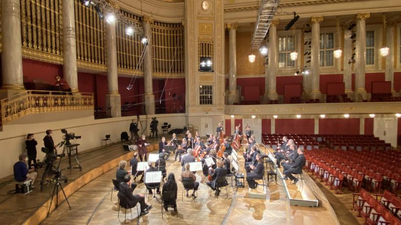 Blick auf ein Orchester in Probe in einem eleganten Konzertsaal mit hohen Säulen, goldverzierten Geländern und roten Sitzen, umgeben von professioneller Filmausrüstung.